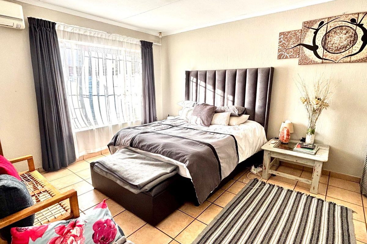 A well-lit bedroom featuring tiled flooring, a wall-mounted air conditioning unit, and large windows with security bars.