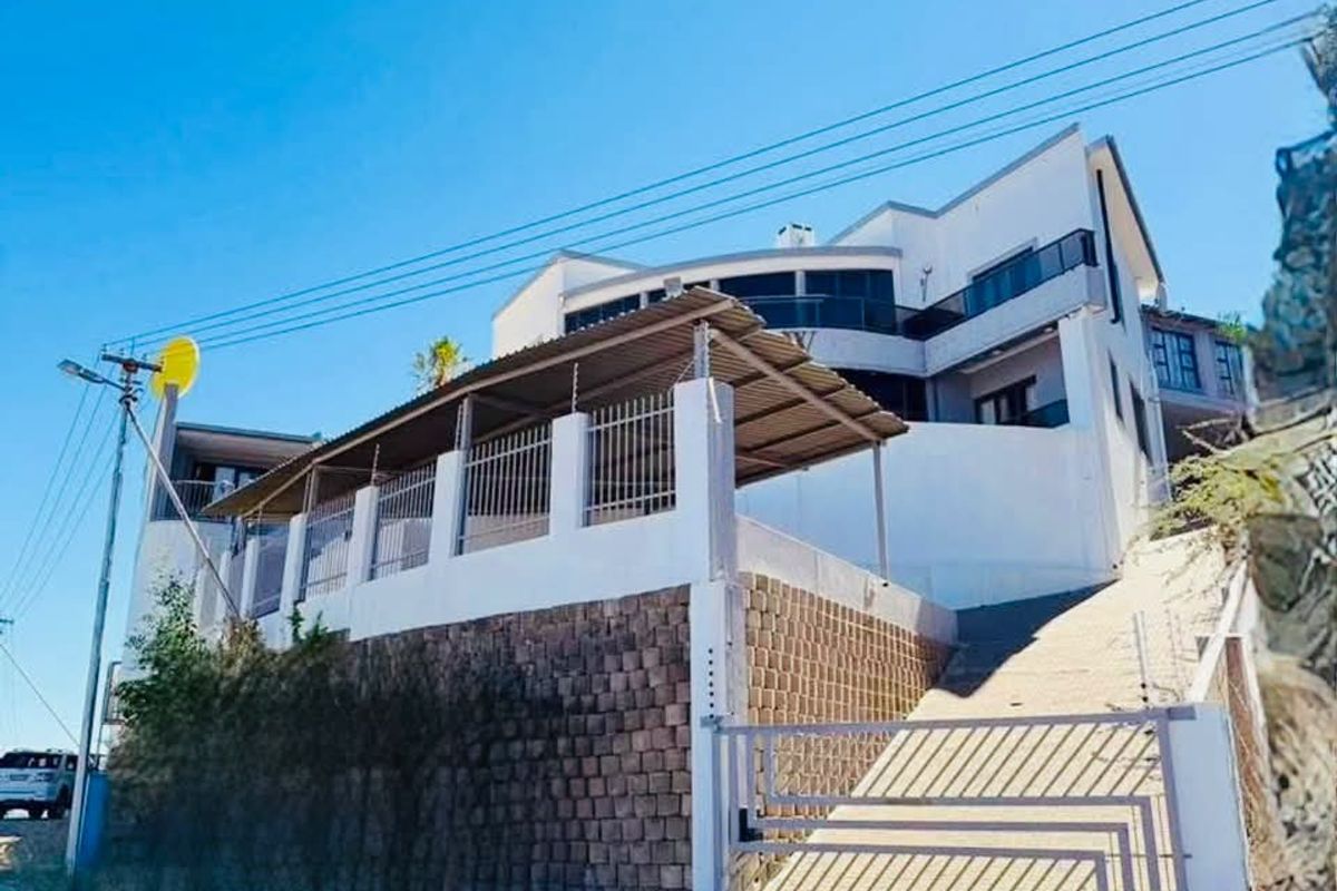 Front exterior view of a multi-level hotel property featuring a paved driveway, metal security gate, retaining wall, and covered carport area.