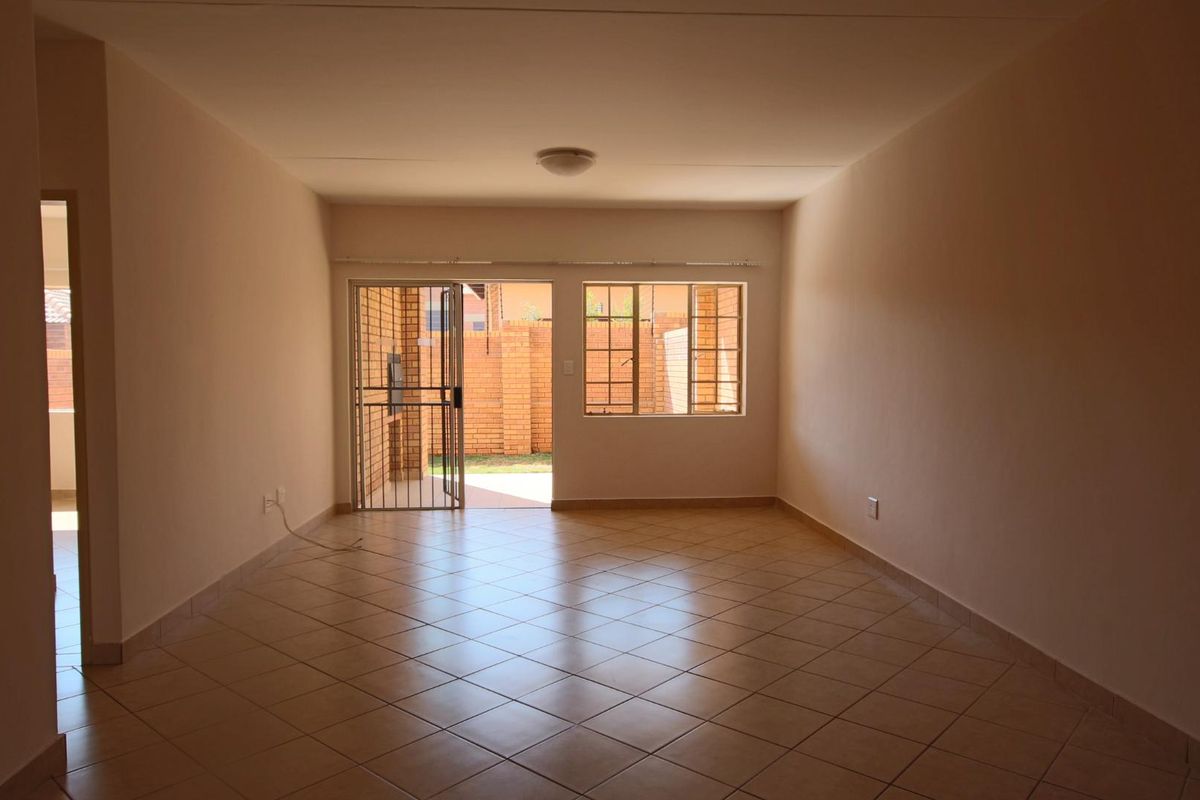 A spacious, empty living area featuring neutral tiled flooring, natural light, and direct access to an outdoor patio through a security gate.