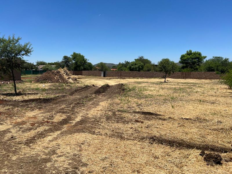 A large, undeveloped plot of land featuring dry grass, scattered trees, and a perimeter brick boundary wall.