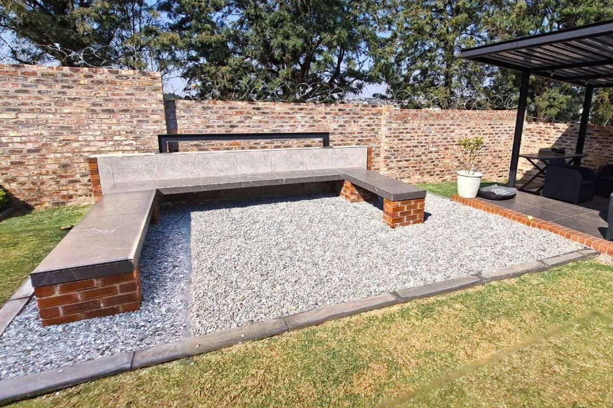A well-maintained rear garden featuring a built-in stone bench, gravel seating area, and a covered patio space enclosed by a brick boundary wall.