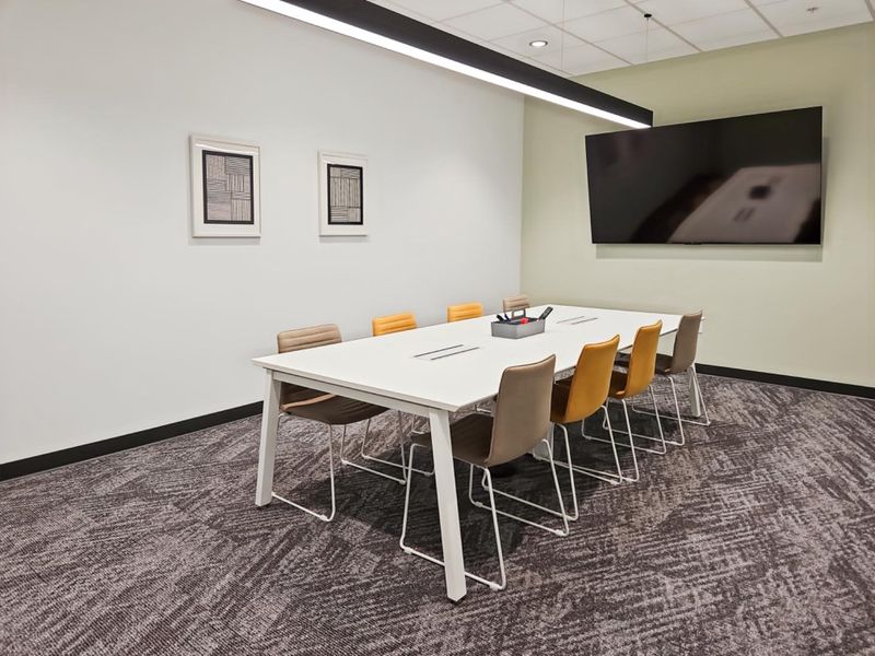 A modern, well-maintained office meeting room featuring carpeted flooring, a suspended ceiling with integrated lighting, and a wall-mounted display screen.