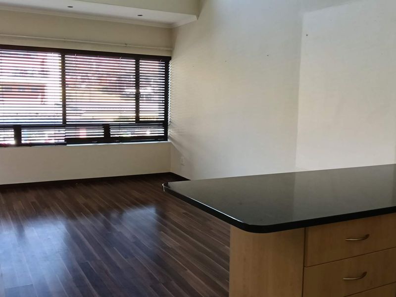 An open-plan living and kitchen area featuring dark laminate flooring, modern veneer cabinetry with black stone countertops, and large windows with venetian blinds.