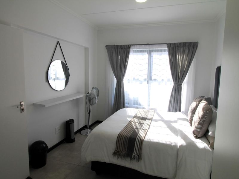 A bright and tidy bedroom featuring neutral tiled flooring, a built-in floating shelf, and a large window with curtains allowing for natural light.