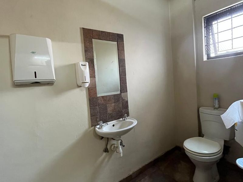 A functional commercial bathroom featuring a wall-mounted basin, tiled mirror surround, and standard toilet with tiled flooring and security window grille.