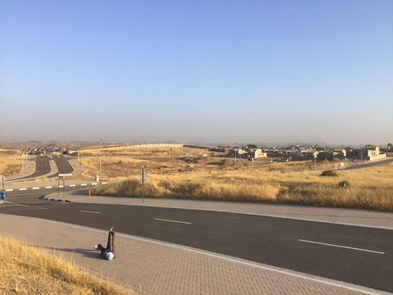 A view of a paved residential street and sidewalk in a developing area with surrounding vacant land and distant housing.