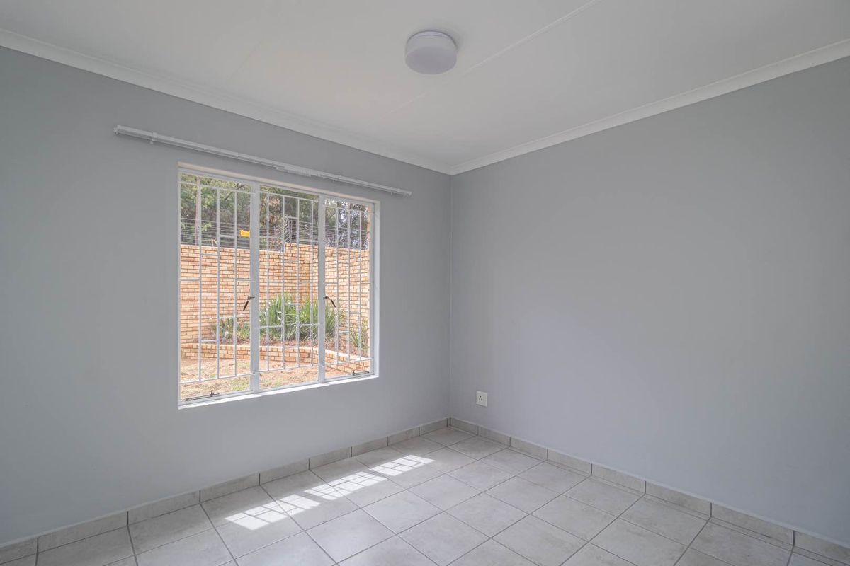 A clean, empty bedroom featuring neutral grey walls, tiled flooring, and a window fitted with security bars overlooking the rear garden area.