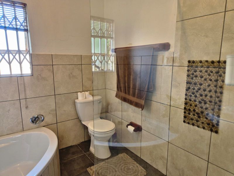 A functional bathroom featuring a bathtub, toilet, and glass shower screen with neutral ceramic tiling and security bars on the window.