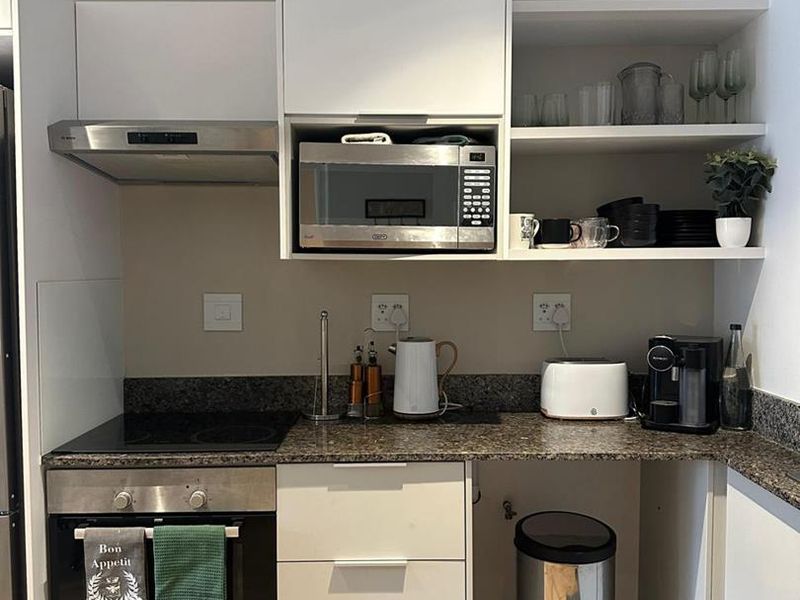 Compact modern kitchen featuring white cabinetry, granite countertops, built-in oven, electric hob, extractor fan, and integrated microwave with open shelving storage.