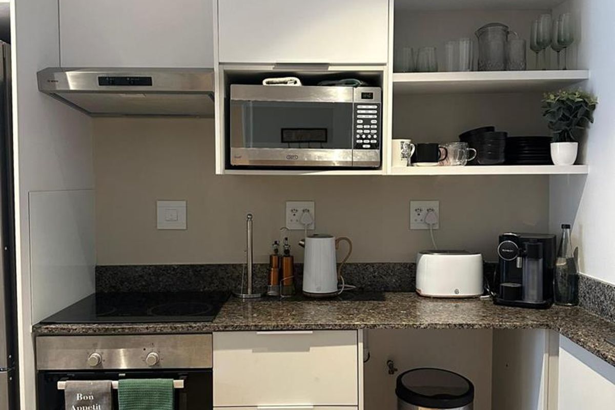 Compact modern kitchen featuring white cabinetry, granite countertops, built-in oven, electric hob, extractor fan, and integrated microwave with open shelving storage.