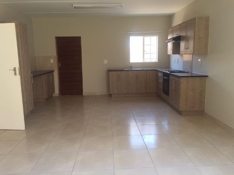 An open-plan kitchen area featuring light wood-finish cabinetry, dark countertops, built-in cooking appliances, and neutral tiled flooring in a clean, vacant space.