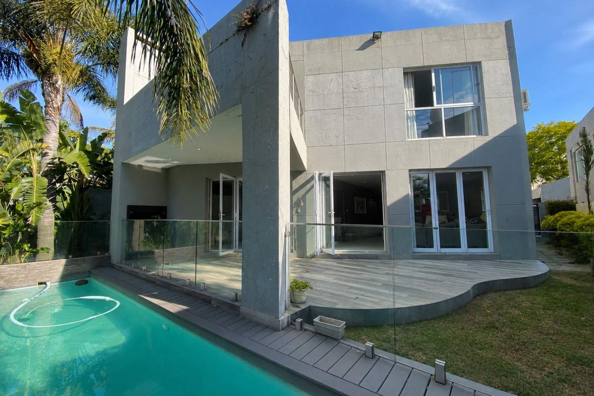 Modern two-story rear elevation featuring a tiled patio, glass balustrades, and a swimming pool set within a landscaped garden.