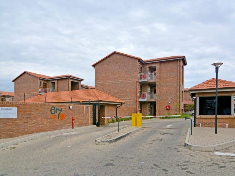 The entrance to the apartment complex features a secure boom gate, guard house, paved driveway, and multi-story brick buildings with tiled roofs.