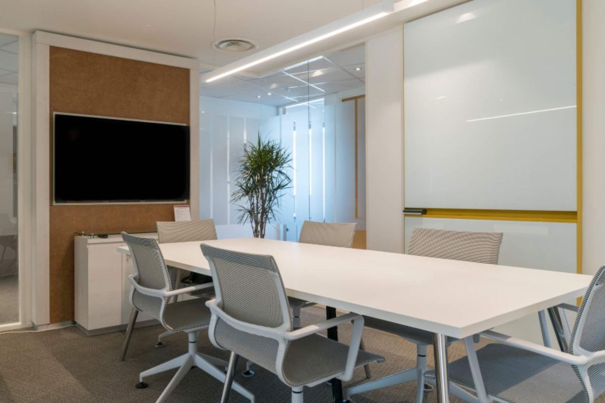 A modern, well-lit office meeting room featuring a glass partition, built-in whiteboard, and cork wall detailing, suitable for professional collaboration.