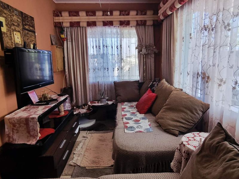 A compact living room featuring large windows with decorative curtains and integrated security bars, providing a functional and lived-in space.