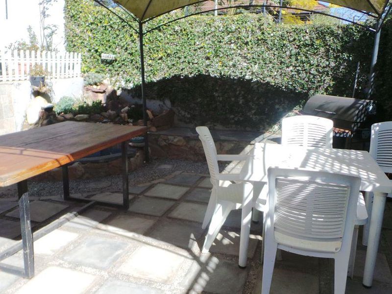 A paved outdoor patio area featuring a fixed gazebo structure, retaining wall, and established boundary greenery, providing a functional space for outdoor dining.