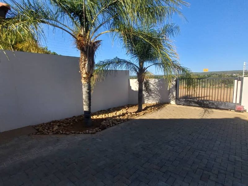 A paved front driveway area featuring a secure metal sliding gate, perimeter walling, and established palm trees.