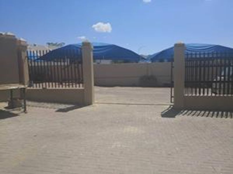 Front exterior view of the industrial property featuring a paved driveway, secure perimeter wall with metal gate, and shade cloth carport structures.