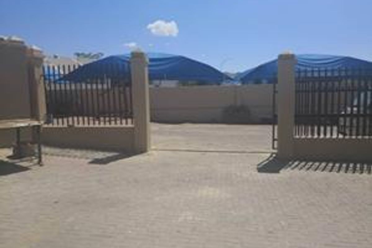 Front exterior view of the industrial property featuring a paved driveway, secure perimeter wall with metal gate, and shade cloth carport structures.