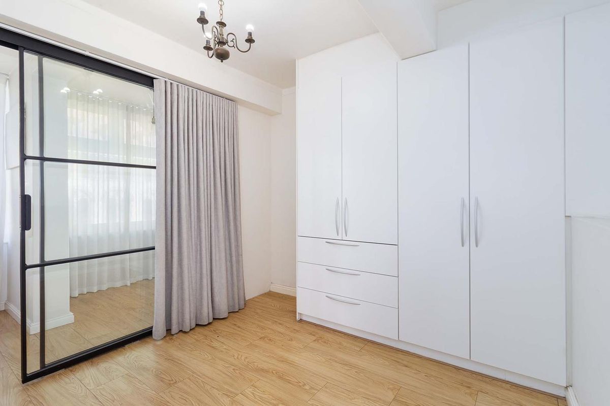 A bright bedroom featuring modern built-in wardrobes, light wood-look flooring, and a stylish black-framed sliding glass door partition.
