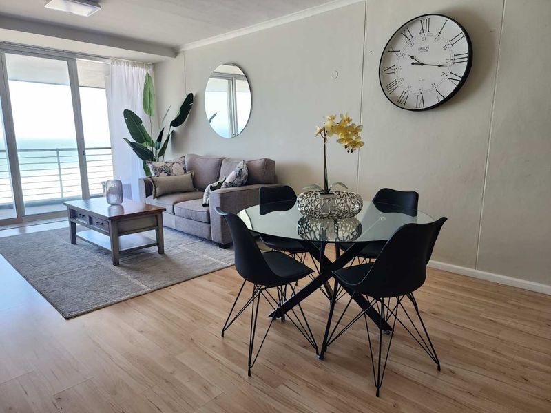 Bright open-plan living and dining area featuring light laminate flooring and large sliding glass doors leading to a balcony with ocean views.