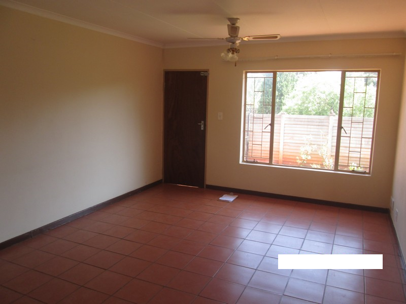 An empty living room featuring terracotta tiled flooring, a ceiling fan, and large windows with security bars, offering a blank canvas for personalization.
