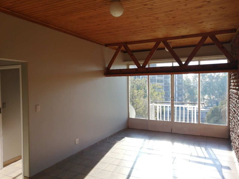 An empty living room featuring a timber ceiling, exposed wooden truss, tiled floors, and an exposed brick wall with access to a balcony.
