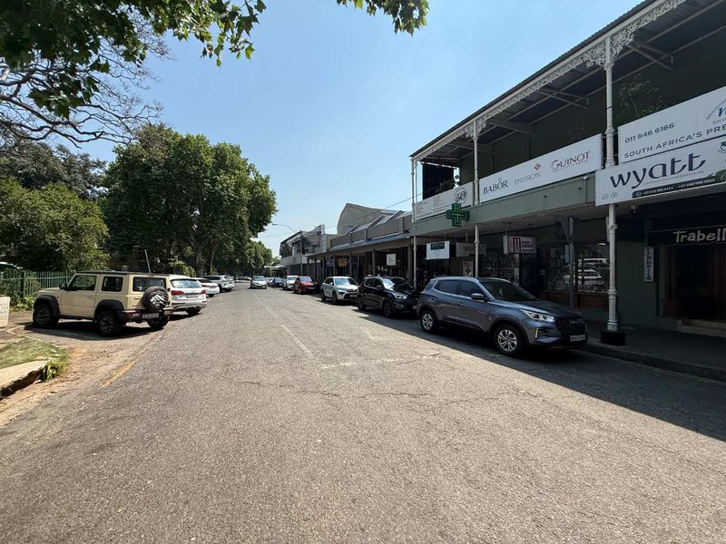A street-level view of a retail strip with storefronts, covered walkways, and on-street parking in a commercial area.