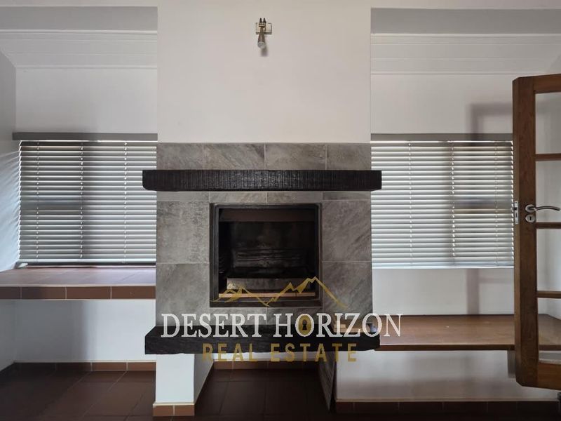A bright living room featuring a central stone fireplace with a timber mantelpiece, tiled flooring, and venetian blinds.