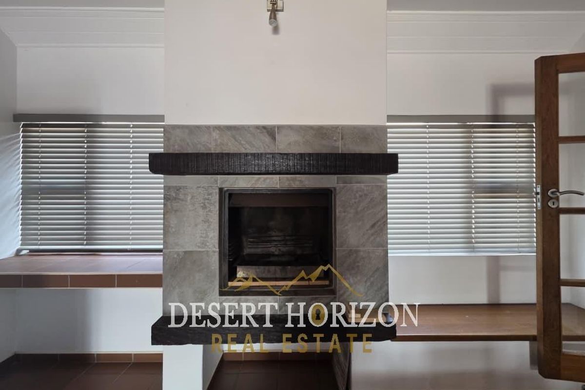 A bright living room featuring a central stone fireplace with a timber mantelpiece, tiled flooring, and venetian blinds.