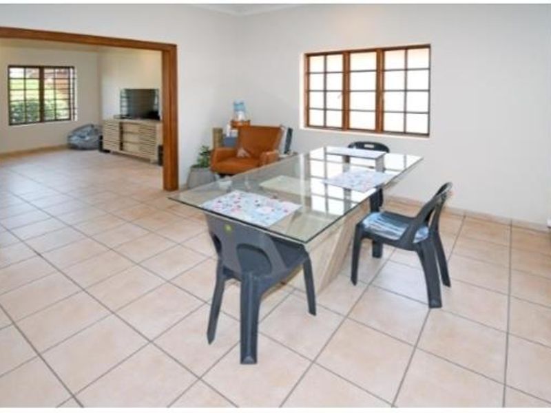 A dining room featuring neutral tiled flooring and wooden window frames, leading into an adjacent living area through a wide wooden doorway.