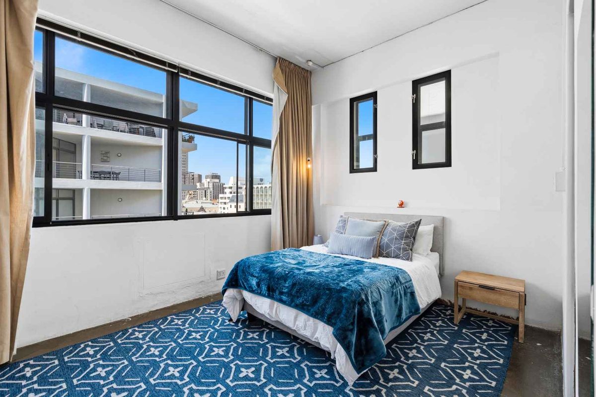 A bright bedroom featuring large black-framed windows with city views, exposed concrete flooring, and built-in wall niches.