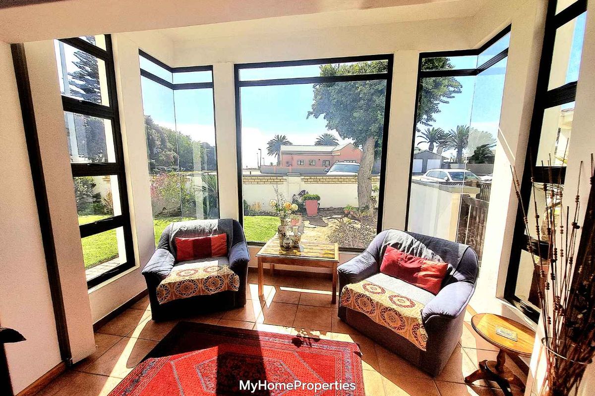 A sun-filled living area featuring extensive floor-to-ceiling glass windows and tiled flooring, offering a bright and airy space with views of the garden.