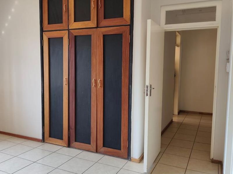 A bright, empty bedroom featuring tiled flooring, built-in wooden cupboards, and a doorway leading to a hallway.