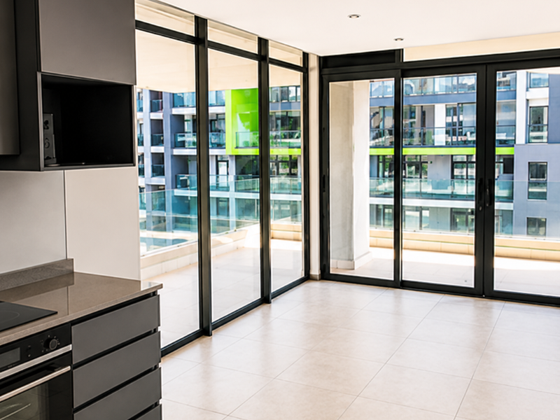 Modern open-plan kitchen and living area featuring sleek dark cabinetry, integrated appliances, large format floor tiles, and expansive sliding doors leading to a balcony.