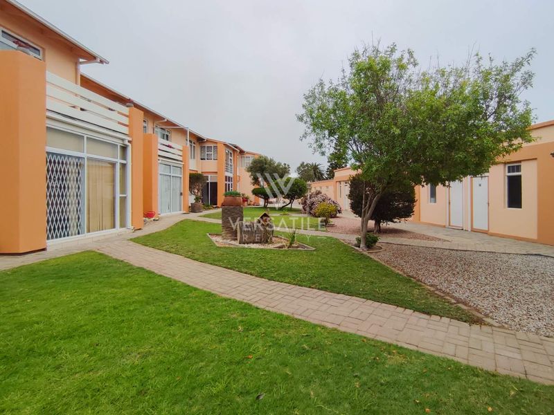 A well-maintained communal garden area featuring paved walkways and access to residential units with sliding glass doors and security gates.
