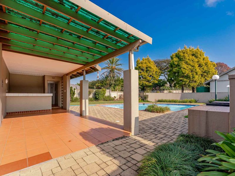 Outdoor entertainment area featuring a covered patio with a built-in bar counter, overlooking a swimming pool and paved garden space.