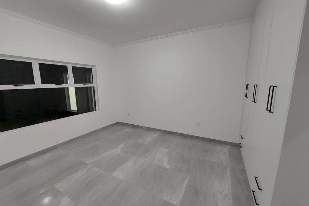 A clean, empty bedroom featuring modern grey tiled flooring, white walls, and ample built-in cupboard storage with contemporary black handles.