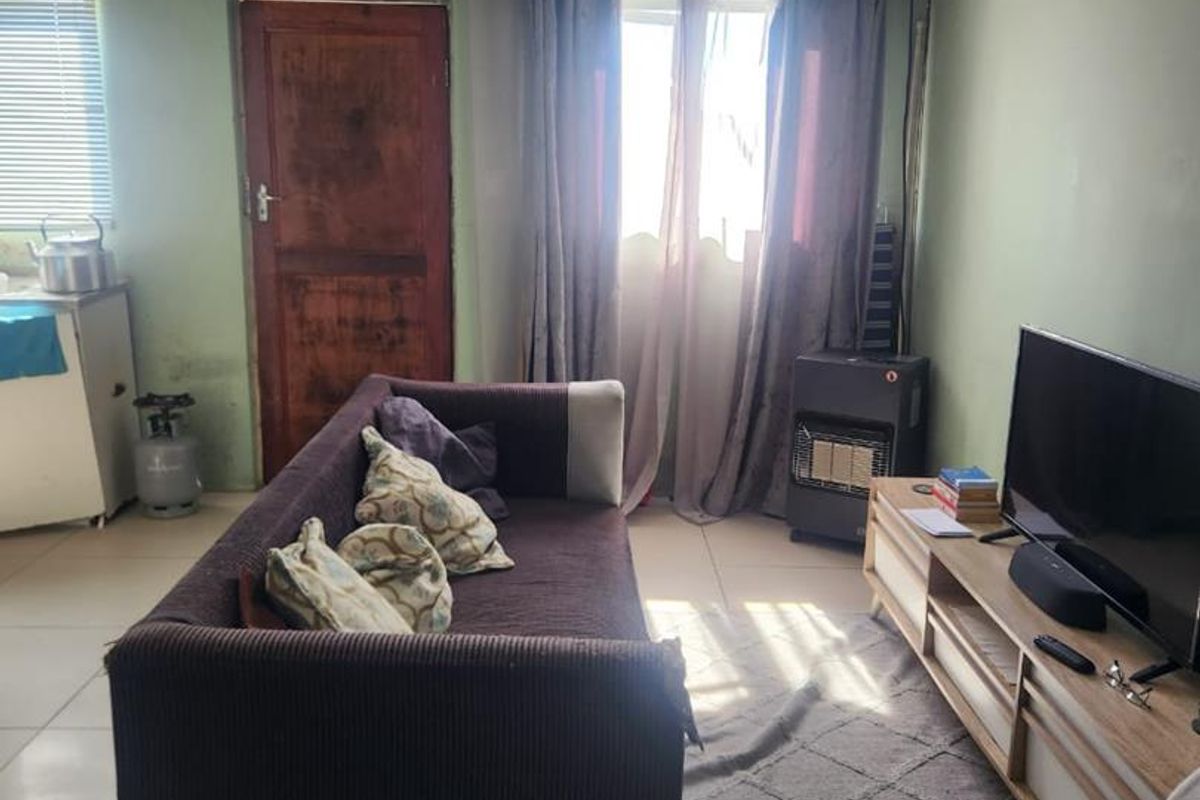 A living room featuring tiled flooring, decorative ceiling panels, and a timber door, showing signs of wear and wall staining.