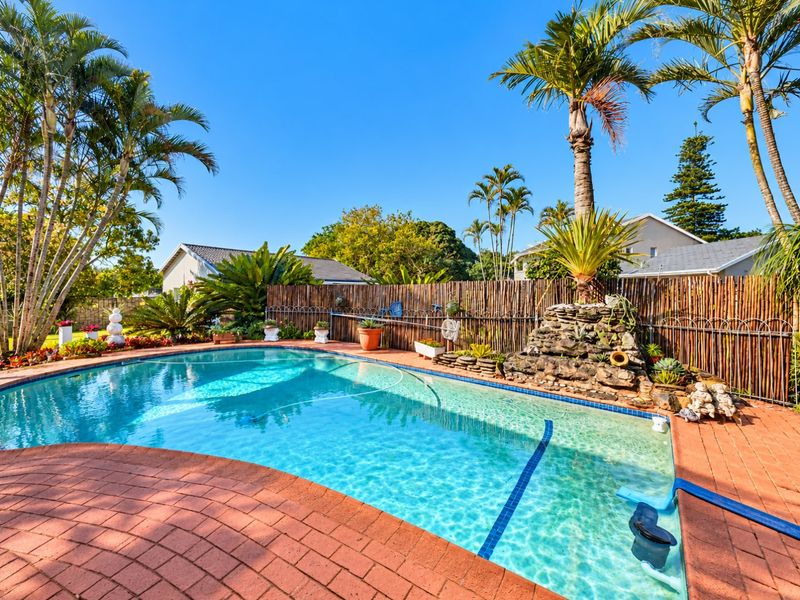 A well-maintained outdoor pool area featuring brick paving, a decorative rock water feature, and established palm trees enclosed by bamboo fencing.