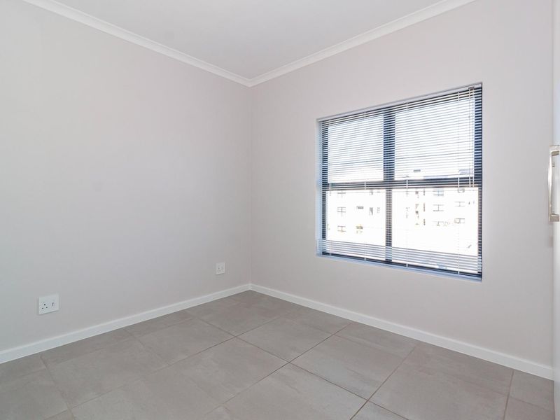 A clean, empty bedroom featuring neutral wall paint, large format floor tiles, and a window fitted with venetian blinds.