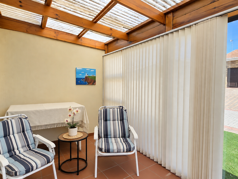 A covered patio area featuring a timber pergola with a translucent roof, tiled flooring, and vertical blinds, providing a comfortable outdoor living space.