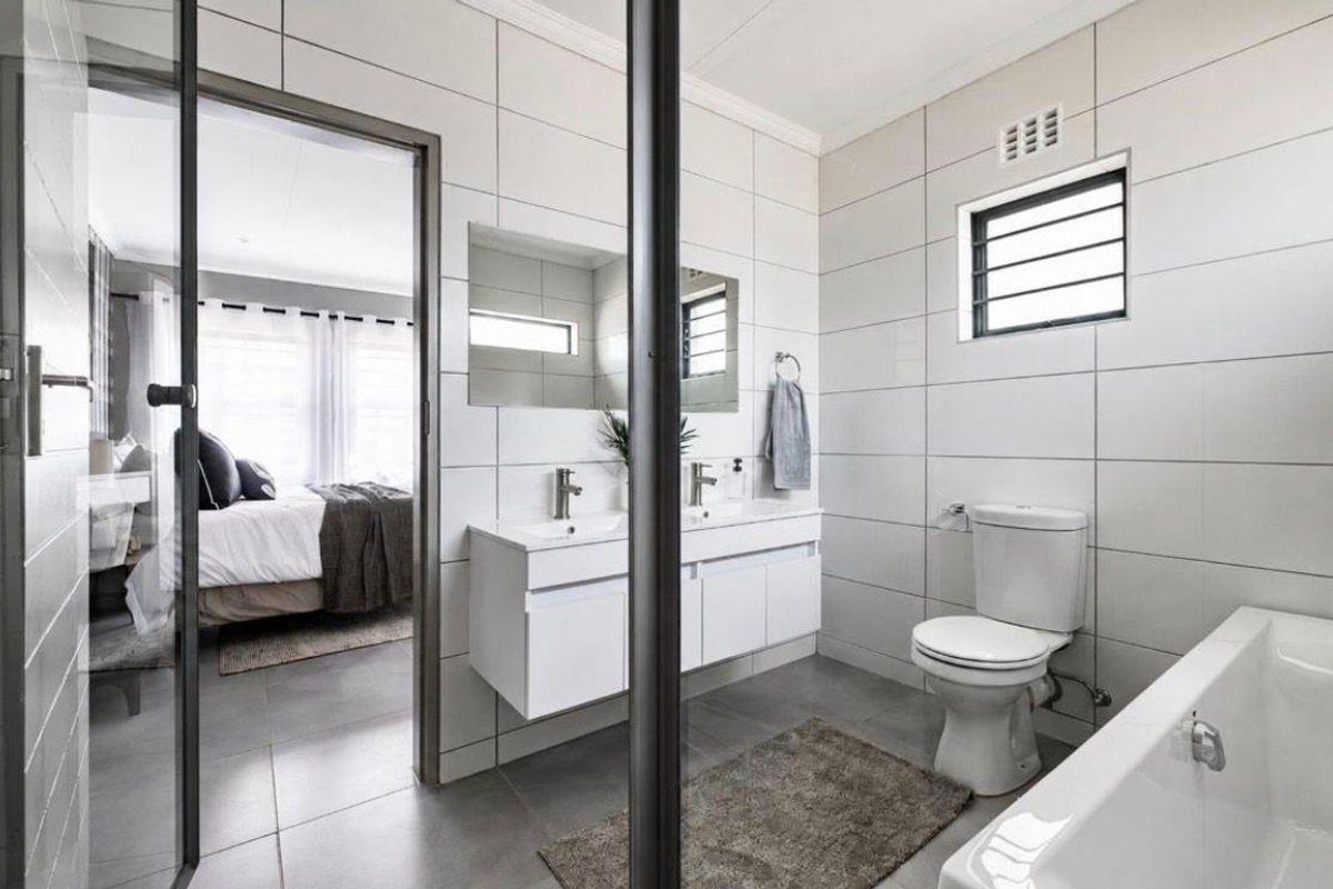 Modern bathroom featuring a double vanity with integrated sinks, a white bathtub, and neutral wall tiling, with an adjacent view into a bedroom.