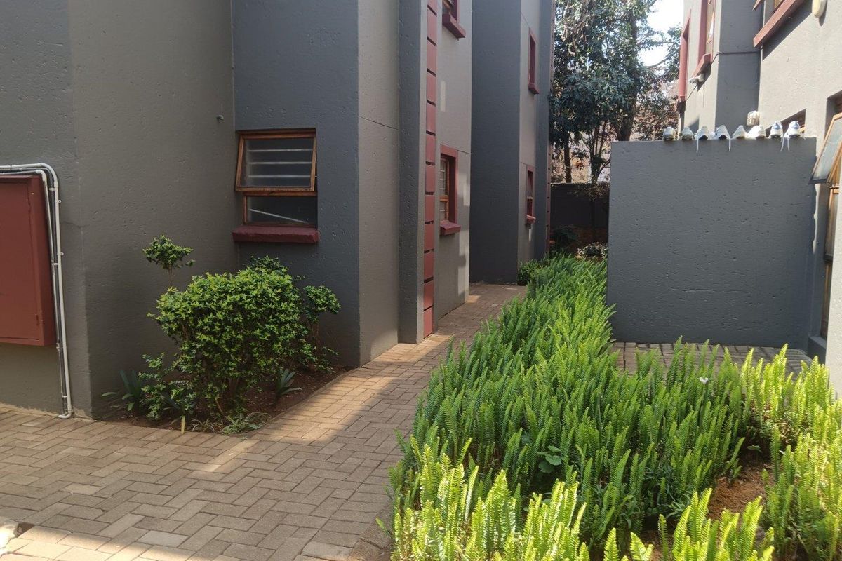 A view of the apartment complex exterior featuring a paved walkway, landscaped garden beds, and dark grey building facades with louvre windows.