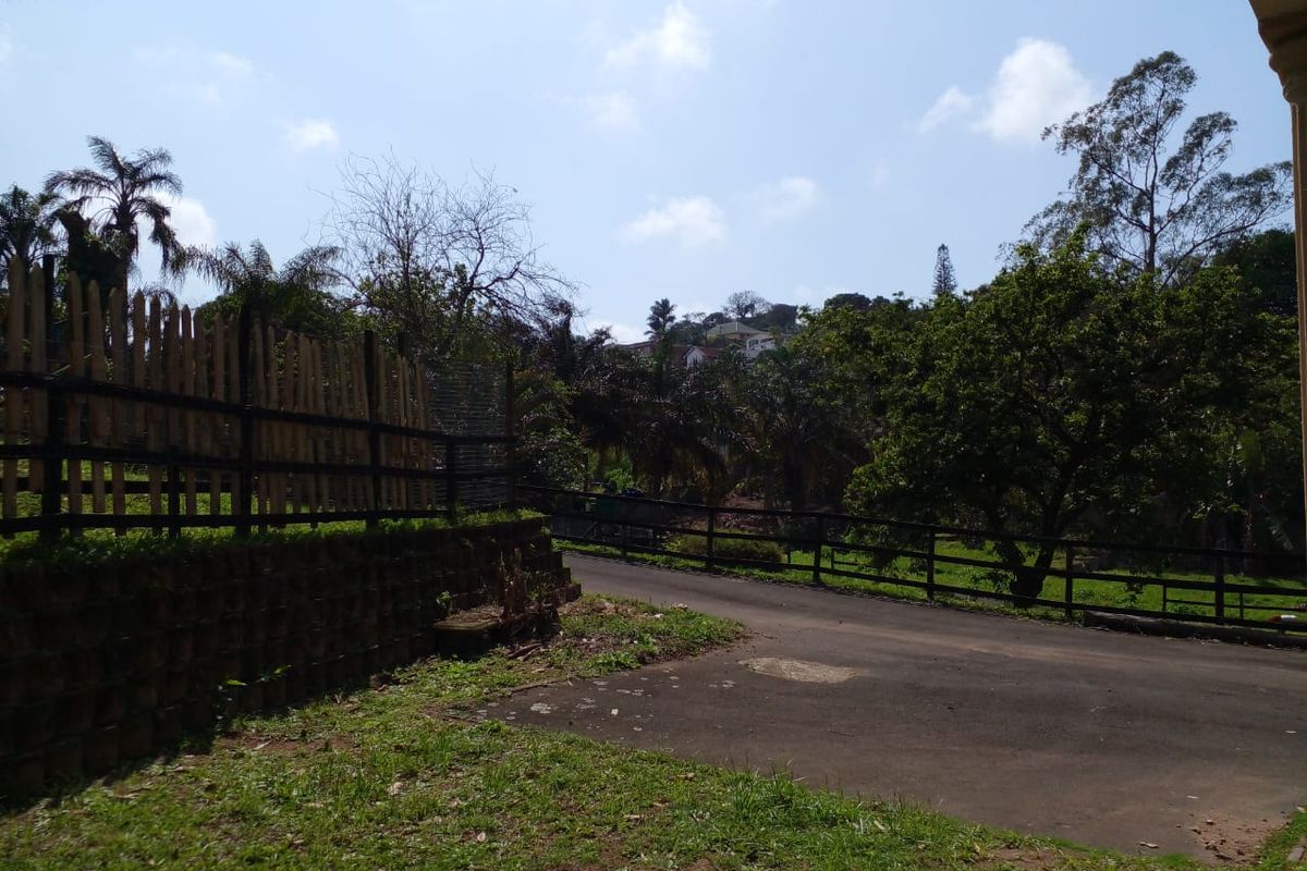 A view of the rear property grounds featuring a paved driveway, timber fencing, and a retaining wall set against a landscaped garden area.