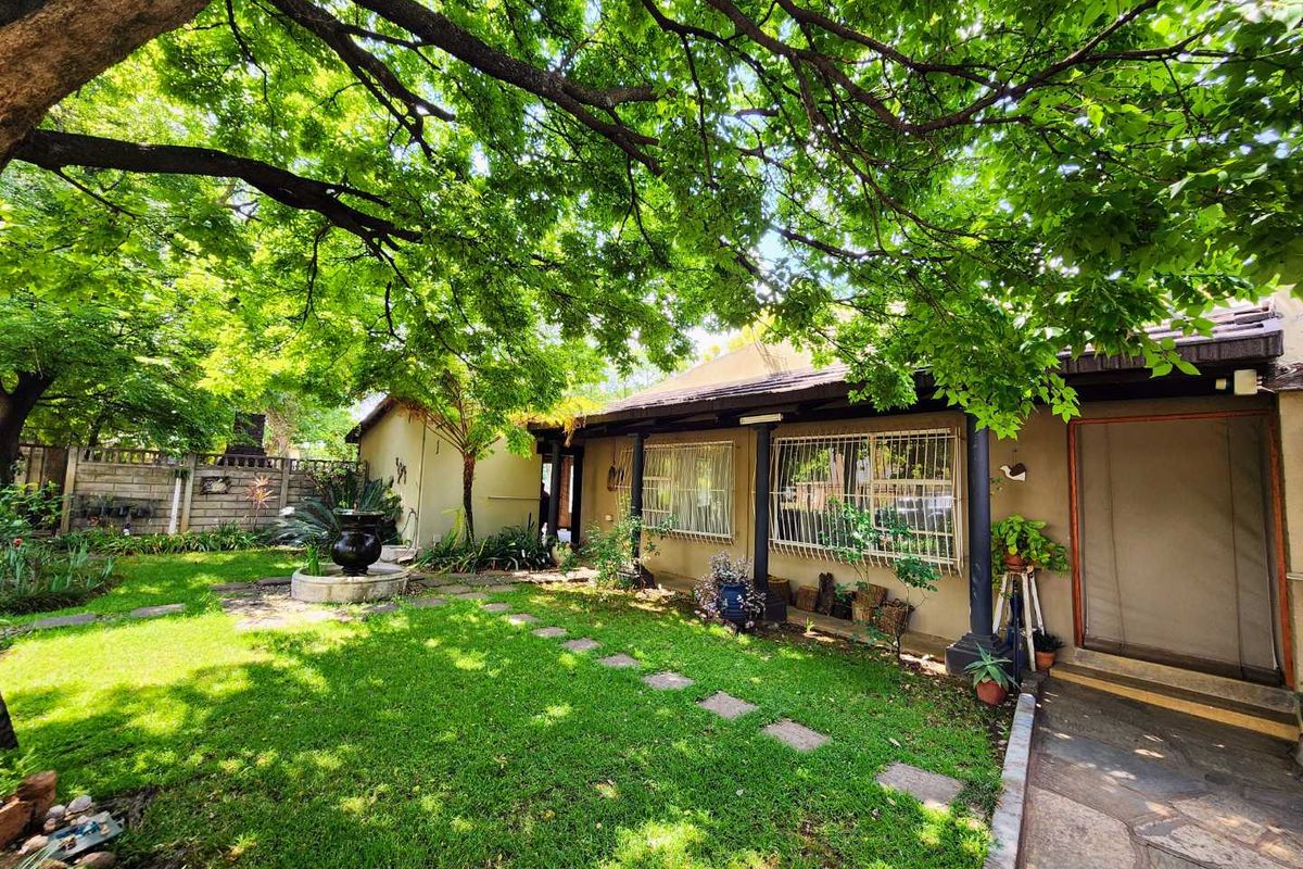 A well-maintained rear garden featuring a lush lawn, mature trees, a stone paved walkway, and a covered patio area with security window bars.