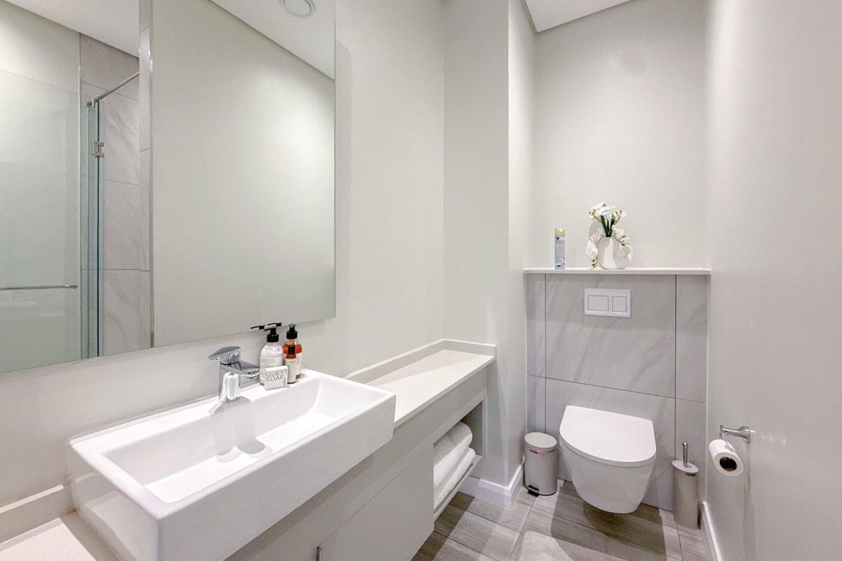 Modern bathroom featuring a wall-mounted toilet, sleek vanity with integrated basin, large mirror, and a glass-enclosed shower with neutral tile finishes.