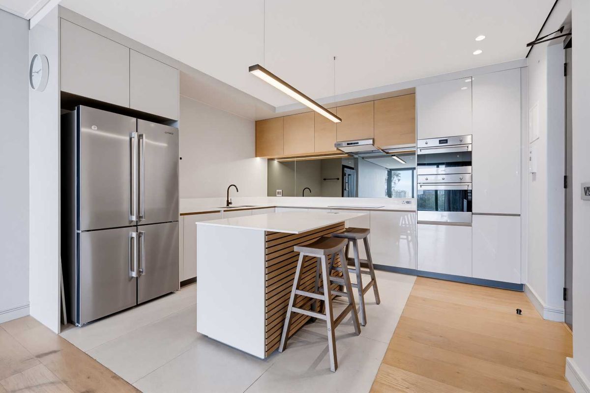 Modern kitchen featuring sleek white cabinetry, timber-accented island, stone countertops, integrated appliances, and a stylish mirror splashback in a contemporary apartment setting.