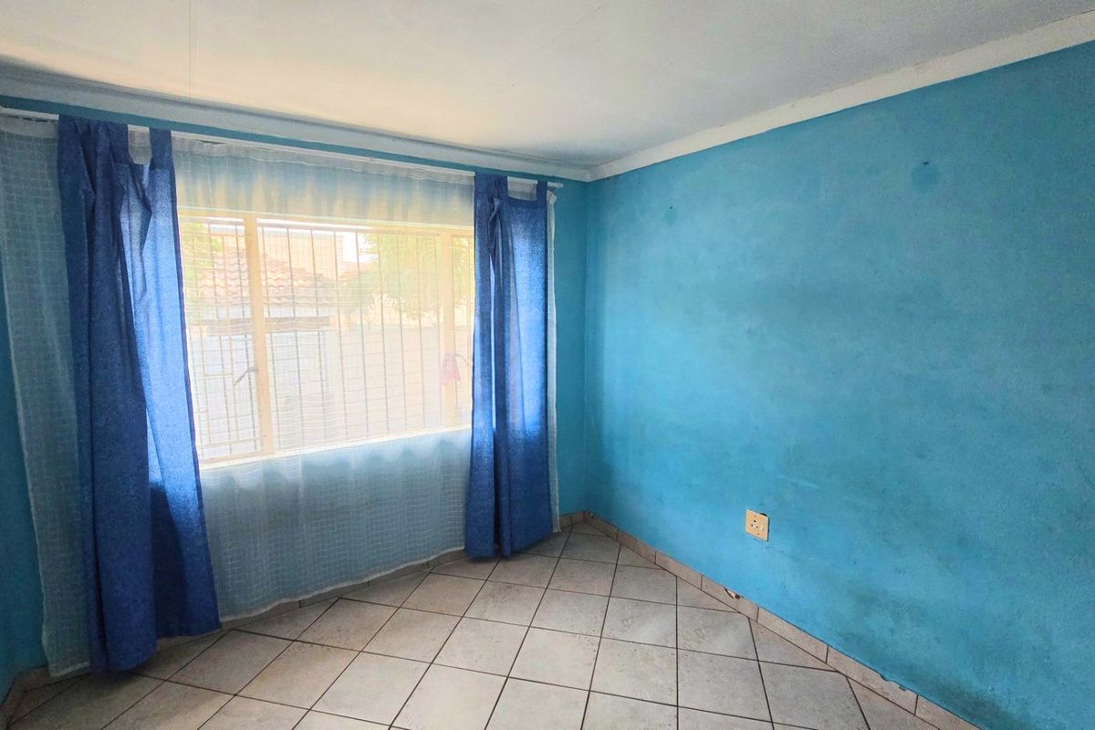 An empty bedroom featuring tiled flooring, security bars on the window, and blue painted walls in need of refreshing.