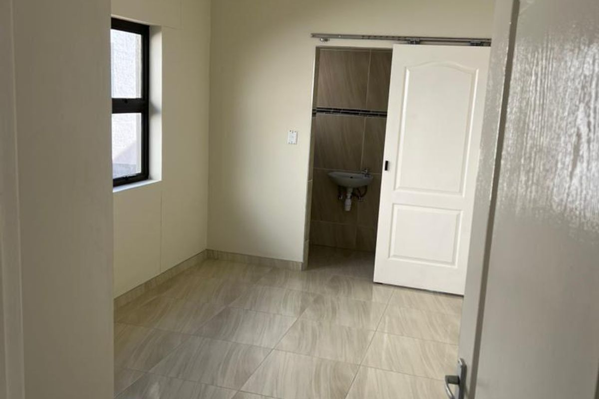 A bright bedroom featuring neutral tiled flooring, a ceiling-mounted light fixture, and an en-suite bathroom area accessed via a sliding door.